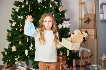 A girl stands in the background of a Christmas tree. The girl is holding a gift. The girl received a medical mask as a gift.