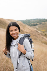 Cheerful African American hiker with backpack looking at camera with blurred landscape at background