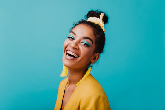 Romantic african young lady with blue makeup laughing to camera. Close-up portrait of cheerful black lady in yellow jacket.