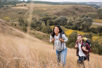 Interracial couple with backpacks walking on hill with grass during trip