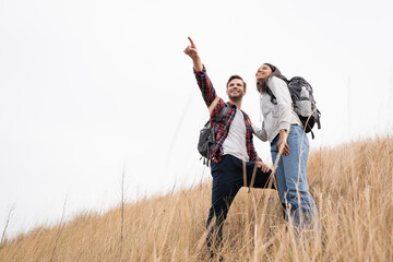 Fototapeta na wymiar Low angle view of smiling man with backpack pointing with finger away near african american woman while standing on grassy hill with sky at background