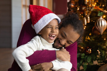 Close up African American man hugging adorable son wearing festive red cap, family celebrating Christmas, sitting near decorated tree at home, loving dad and little child boy cuddling, winter holiday
