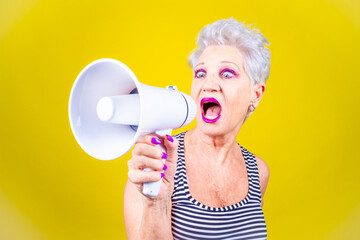 Senior grey haired woman screaming loudly in a megaphone on yellow background demonstrating