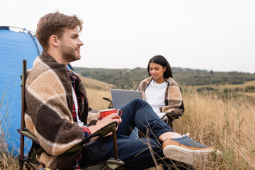 Man wrapped in blanket holding cup near african american girlfriend using laptop on lawn on blurred background