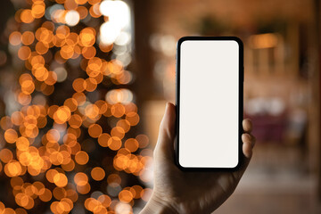 Close up smartphone with empty screen mockup on holiday background with Christmas tree, customer holding phone in hand, shopping online, purchasing gifts, searching information, browsing apps