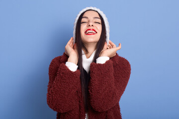 Dark haired girl puts on winter hat over isolated blue background laughing, showing her teeth, wears red lipstick, expressing positive emotions, dresses faux fur coat.