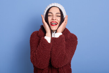 Positive lovely woman with red lips, keeps both hands on cheeks, wears white winter sweater and warm coat, looks at camera with charming smile, isolated over blue wall, lady laughing happily.