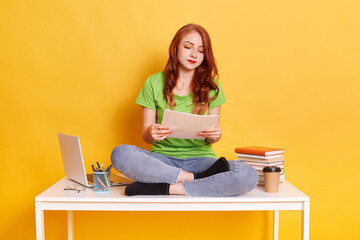 Young woman works at home, holding paper documents in hands and reading it wit concentrated look,...