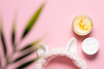 Spa and wellness concept. Funny hair band, natural facial scrub and moisturizing cream on pink pastel background with blurred palm leaf. Natural cosmetic and face care concept.