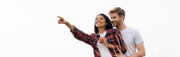 Smiling man hugging african american girlfriend with cup near grass isolated on white, banner