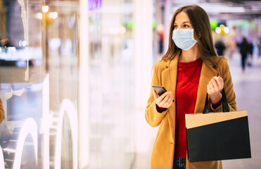 Confident trendy woman in a safety medical mask with a shopping bag and smart phone is walking in the mall during black friday