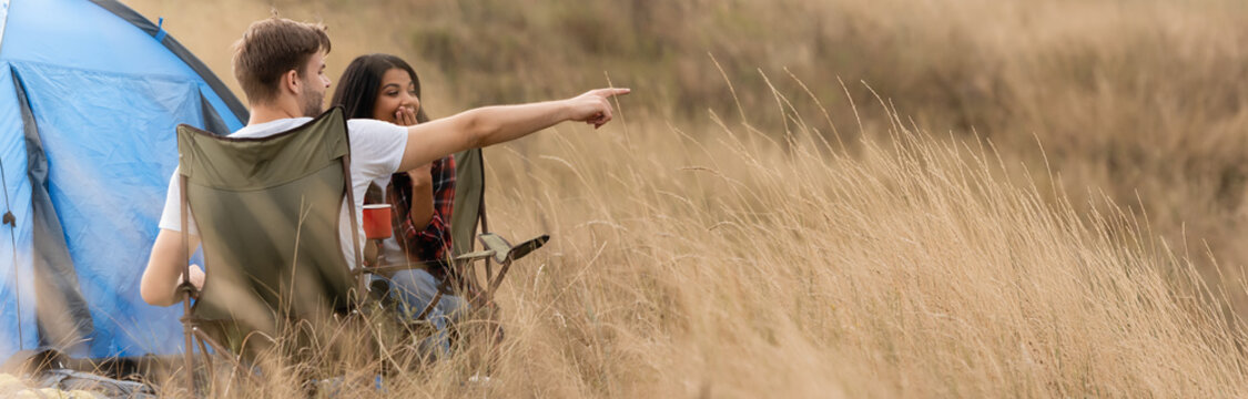 Man Pointing With Finger Near African American Girlfriend With Cup During Camping On Lawn, Banner