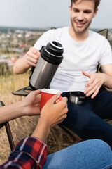 Naklejka premium African american woman holding cup near smiling man with thermos on blurred foreground during camping