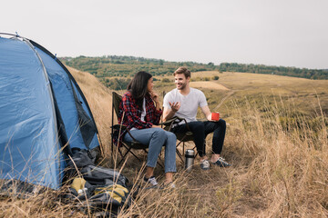 Smiling man with cup talking to african american girlfriend near backpacks and tent on meadow