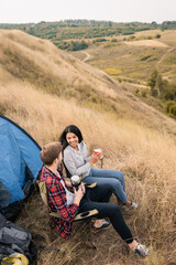 Fototapeta na wymiar Smiling interracial couple holding cup and thermos near tent during camping in field