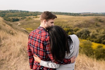 Back view of young man hugging african american girlfriend with grassy hills on blurred background