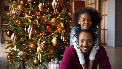 Head shot portrait smiling African American adorable girl hugging father from back near festive...