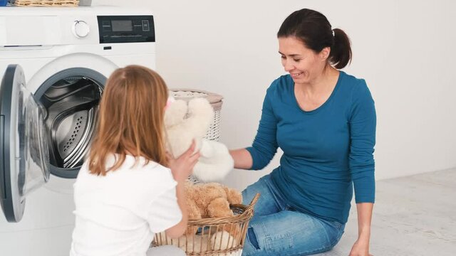 Mother and little daughter loading plush toys into washing machine in laundry room