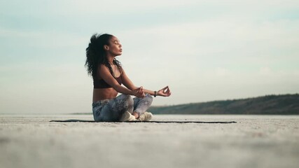 A calm african american woman wearing sportswear is doing yoga sitting on the mat outside at the beach - Powered by Adobe