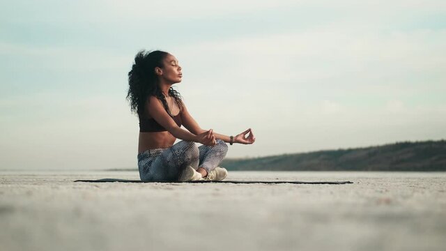 A calm african american woman wearing sportswear is doing yoga sitting on the mat outside at the beach