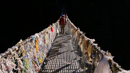 Group of trekkers crossing the popular Hillary suspension bridge decorated with colorful Buddhist prayer flags in the midday sun with hardly visible blacked background near Namche Bazar, Nepal.
