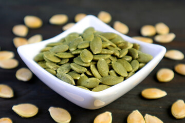 Closeup a bowl of roasted pumpkin seeds with raw seeds scattered around on wooden background