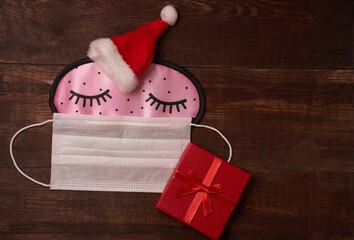 Sleep mask and protective mask Santa's cap sleeps with gift box, flatlay with space for text on brown wooden background