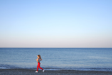 Positive sportive woman on a summer morning jogging on the beach in red leggings on the background of the sea coast