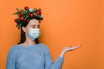 Young Caucasian woman in medical face mask, keeps hand raised, dressed in winter sweater and...