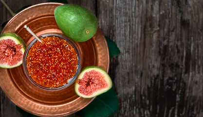 Fig jam in a glass jar on a copper Turkish dish, with halves of fresh figs and a fig leaf next to it. Top view, delicious fig dessert. Dark wooden background with copy space