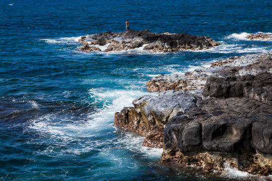 Black laval rocks on a jagged coastline with waves breaking,one person standing on an outcrop.