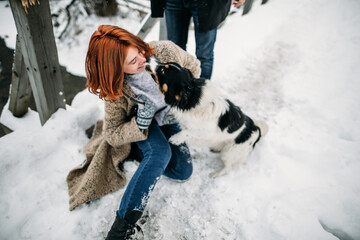A dog stretches to lick a red-haired girl in winter sitting on the snow on christmas