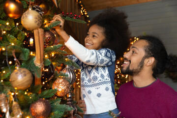 Close up happy African American father with little daughter decorating Christmas tree, smiling dad holding adorable child, helping, family preparing home to New Year celebration, family having fun