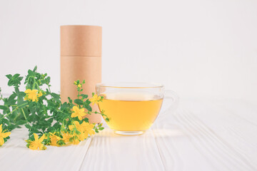 Cup of fresh herbal antioxidant tea, Hypericum flowers and blank paper packaging tube on white table. Alternative medicine, herbalism and healthcare concept. Zero waste tea packaging, mockup image