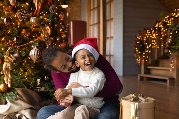 Overjoyed African American man hugging adorable little son wearing festive red cap, family celebrating Christmas, opening gifts, sitting on warm floor at home near tree, enjoying winter holidays