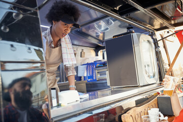 young afro-american female employee in fast food service writing down the order looking at  young caucasian beardy hipster with a cap