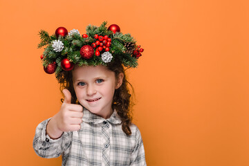 Close up portrait of cute little kid girl showing thumbs up like gesture, wears spruce holiday wreath around head, isolated over orange background studio with copy space. Nappy New Year concept