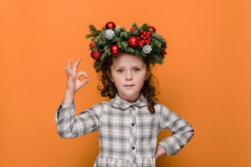 Studio shot of preschooler girl kid in traditional Christmas wreath, makes okay gesture, enjoys life, isolated over orange studio wall with copy space, says ok. Happy New Year merry holiday concept