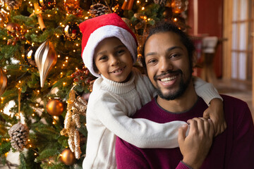 Head shot portrait African American son and father celebrating Christmas, smiling dad piggy backing little boy wearing red cap and warm sweater, looking at camera, sitting near festive tree at home