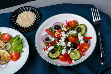 White plate with grilled zucchini with cherry tomatoes on lettuce and Greek salad on a blue napkin with a fork beside sesame seeds.