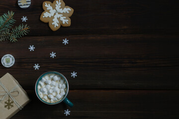 Obraz na płótnie Canvas Christmas New Year card. Cup of hot drink with marshmallows, New Year's gift, gingerbread, snowflakes, candles on a dark wooden background. Festive concept. Top view. Copy space.