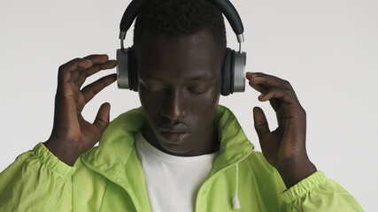 Handsome African American guy dressed in bright jacket listening music in headphones over white background