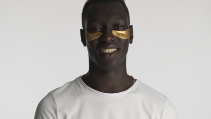 Attractive African American guy with eye patches smiling on camera over white background. Facial treatment