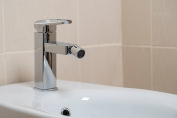 Details of white ceramic bidet with water tap in modern bathroom with beige tiles background. Close...