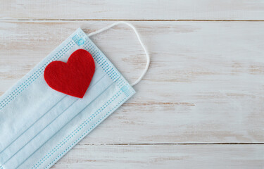 A felt heart lies on a medical protective mask on a wooden white background. Copy space