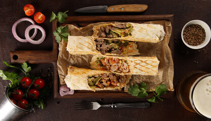 Shawarma with meat and vegetables. Flatbread with lamb, chicken, pickles, onions, mustard and sauce on a wooden board, beer and spices. Fork, knife, herbs, cherry tomatoes. Background image, top view