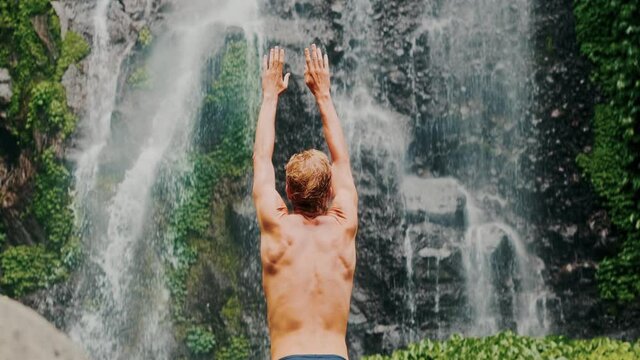 Back view of happy young strong man standing by waterfalls with arms outstretched, feeling freedom and power of nature. Man at waterfall raising his hands in feeling closer to nature. Travel to Bali.