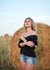 Young blond woman, wearing black jacket and jeans shorts, standing by wheat bale on rural field on summer evening. Three-quarter female portrait during sunset at natural landscape.