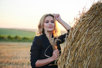 Young blond woman, wearing black jacket and jeans shorts, leaning on wheat bale on rural field on summer evening. Three-quarter female portrait during sunset at natural landscape.