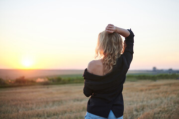 Young blond woman, wearing black jacket and jeans shorts, posing with back to camera on wheat field on summer evening. Creative stylish female portrait during sunset at natural rural landscape.